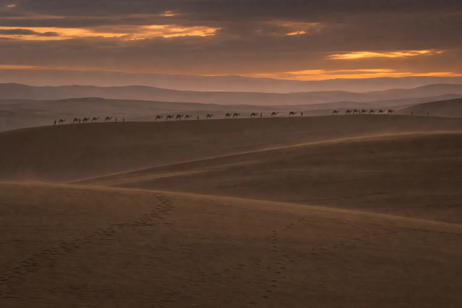 Long distant camel caravan crossing a desert dune ridge beneath orange evening clouds