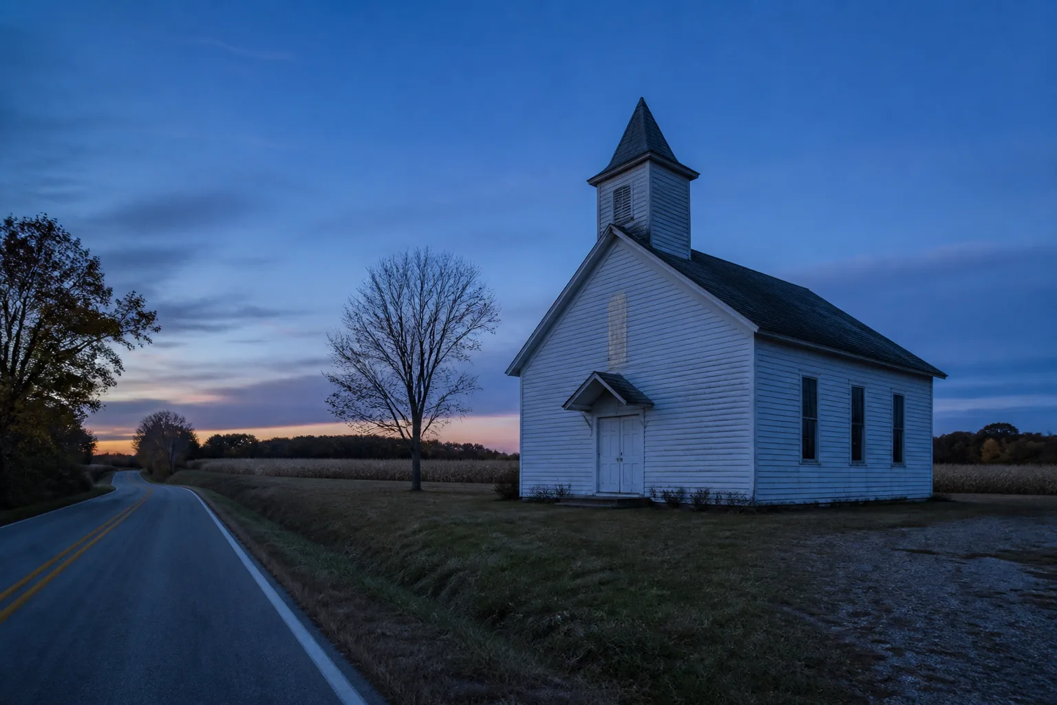 Small white rural church beside an empty road and bare tree at blue hour
