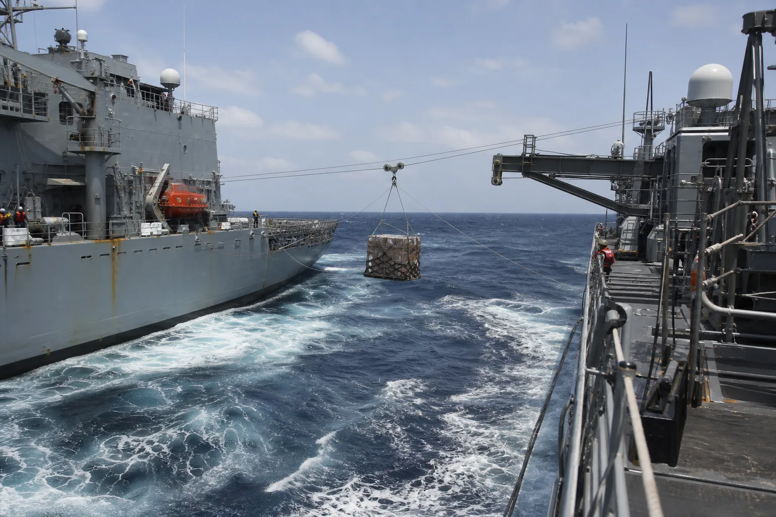 Netted cargo pallet suspended on a highline between two naval ships traveling side by side at sea