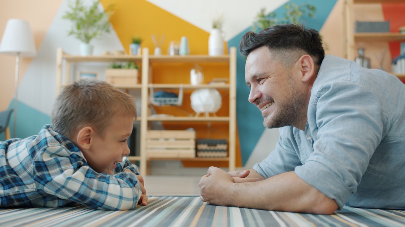 A father and his young son lie on the living-room floor, face to face, smiling at each other.
