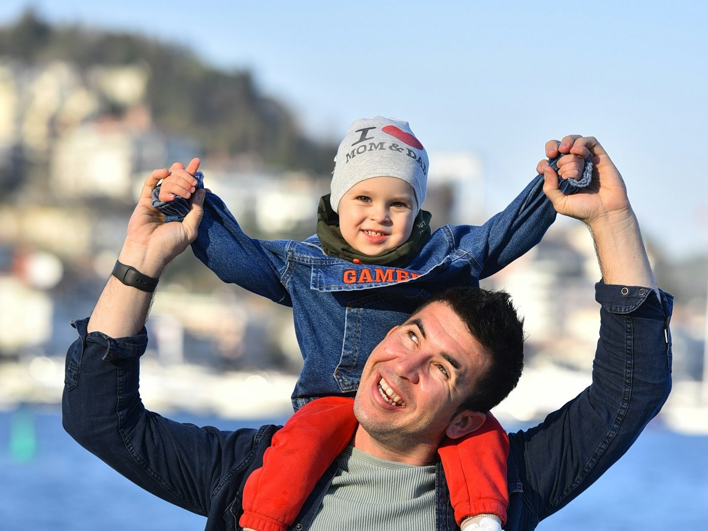 A laughing father carries his small son on his shoulders by the water, holding both of his hands.