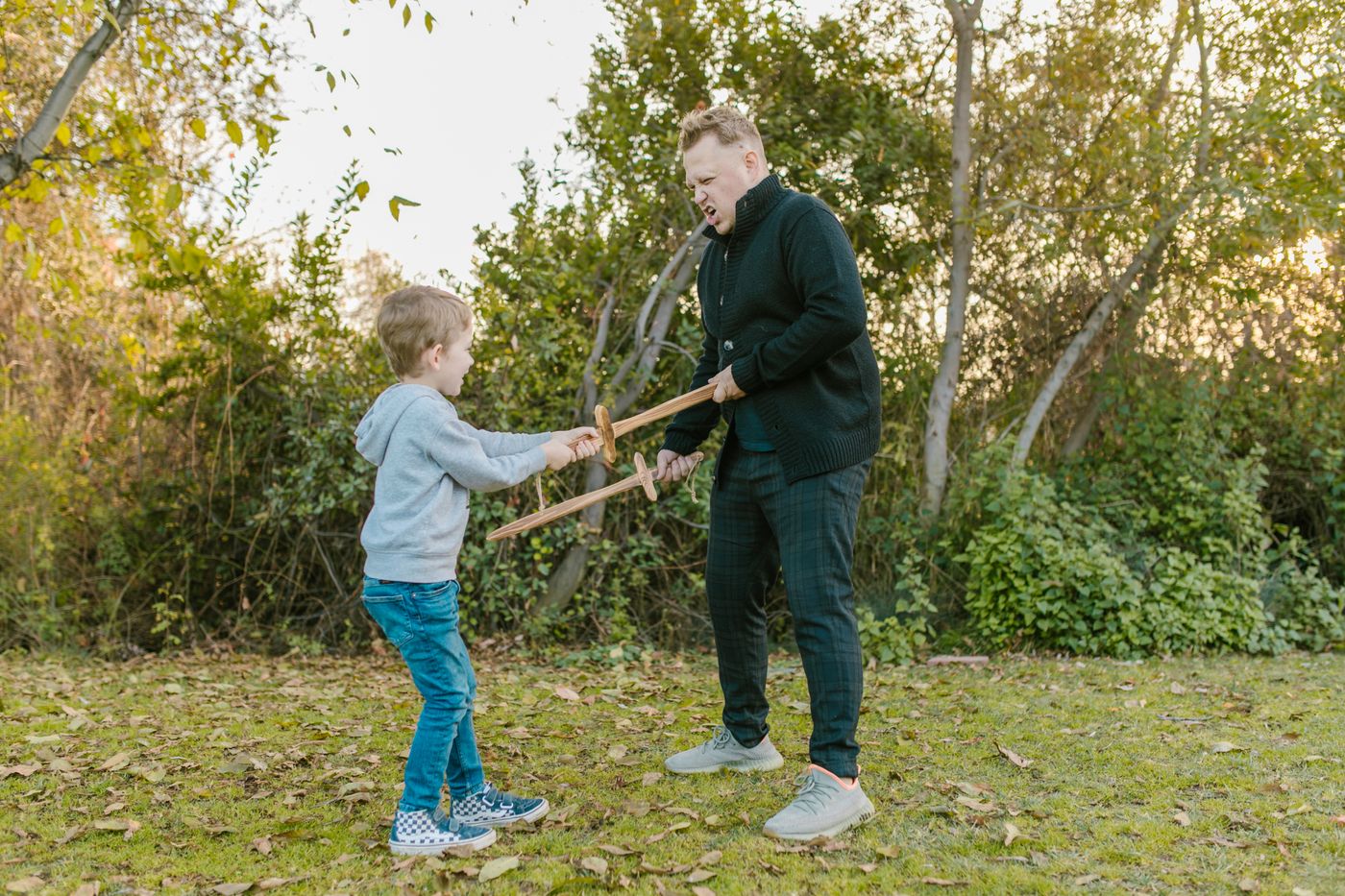 A boy and his father square off with wooden swords in a leafy backyard.